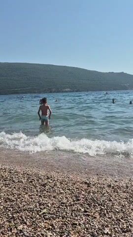 The video shows a young person standing in shallow ocean water with waves gently breaking on a pebble beach in the foreground. The person is facing away from the camera, with their hands on their hips. In the background, several other people are visible in the water, and a green, tree-covered hill rises behind the sea. The sky is clear and blue.