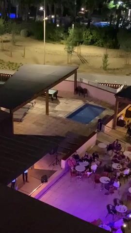 The video shows an aerial view of an outdoor area at night. In the foreground, there is a patio with several round tables and chairs, occupied by groups of people. A stage with musical equipment is set up on the left side of the patio, and purple lighting illuminates the area. In the background, there is a small swimming pool under a shaded canopy, with a pool table nearby. Further in the distance, there is a larger pool area and other outdoor spaces, with trees and lights visible. People are also seen dining and socializing at tables in a covered area to the right of the patio.