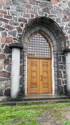 The video shows a close-up view of a stone building, likely a church or cathedral, with an arched entryway. The building's facade is constructed from rough-hewn stones of various colors, including gray, pink, and black. Two large, light-colored stone pillars frame the entrance. Above a set of double wooden doors, there is a large, arched window divided into many small panes by a grid pattern. As the camera pans upwards, more windows and architectural details of the stone tower are visible. The ground in front of the building is covered with green grass and small white flowers. The overall impression is of a historic, sturdy structure.