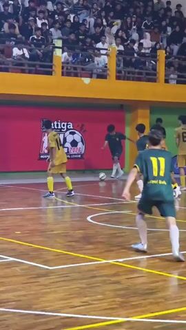 A futsal match is underway. The court is wooden with yellow and white lines. Spectators fill the bleachers in the background. On the court, several young men in athletic uniforms are playing. Two players in green and yellow jerseys are in the foreground, one with the number 11 on his back. Another player in a yellow jersey with the number 14 is in the background. The players are actively engaged in the game, dribbling and kicking the ball. The scene captures a moment of intense play.