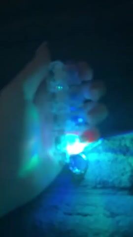 A hand holds a keychain with four clear plastic cubes stacked vertically. Each cube has a small light inside, illuminating in various colors. The lights cycle through blue, green, and red. The background is dark and textured.