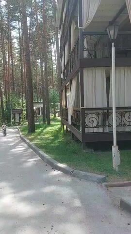 The video pans from right to left, showing a wooden building with curtains on multiple levels. To the left of the building, there is a pathway bordered by grass and tall trees. A person on a bicycle is seen riding on the pathway in the distance, followed by another person who appears to be walking or riding a scooter. Further down the path, a parking lot with several cars is visible, surrounded by grass and trees. The scene appears to be an outdoor recreational area or resort.