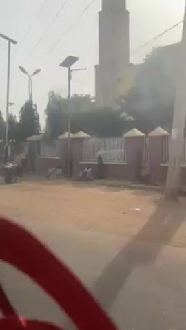 The video, shot from the interior of a moving vehicle, depicts a dusty street scene in an area that appears to be a lower-income neighborhood or possibly a rural area. The visual quality is poor due to dust or haze. In the initial shot, individuals are seen sitting or working near a fence and a building with a tower-like structure. The camera then shifts, revealing more of the street and other buildings, but the hazy conditions make details difficult to discern. The red steering wheel and part of the dashboard are visible inside the vehicle, with a red logo/design on the windshield. The primary action is the movement of the vehicle through the area. There is no visible interaction between the people on the street and the occupants of the vehicle.