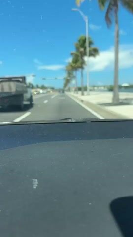 This is a first-person perspective video taken from inside a vehicle, likely a car, looking out through the windshield. The scene is a road lined with palm trees under a bright blue sky with scattered white clouds. The sun appears to be high, suggesting it's daytime, likely midday.

The road ahead shows multiple lanes of traffic. In the left lane, a truck is visible. Further down the road, traffic lights are present, with green lights illuminated, indicating traffic is moving. There are other vehicles in the distance. On the right side of the road, there is a sidewalk and what appears to be a body of water or a very bright, open area, possibly the ocean or a bay, given the presence of palm trees and the likely location. A white sign with the number "35" is visible on the right side of the road, suggesting a speed limit.

The foreground of the video is dominated by the car's dashboard, which is dark gray and textured, with some white dust or smudges visible on its surface. The overall mood is casual and suggests a road trip or a drive through a sunny, tropical location. The blurriness of the background and the movement of the car imply that the vehicle is in motion. The clear weather and vibrant blue sky suggest a pleasant day. The location cues, such as the palm trees, the specific type of road infrastructure (traffic lights suspended over the road), and the speed limit sign, point towards a location in the United States, specifically Keys West, as indicated in the prompt.