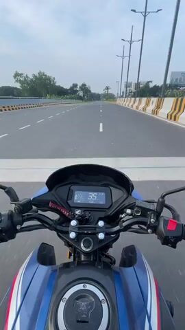 The video provides a first-person perspective of riding a motorcycle on a paved road. The rider's hands are on the handlebars, and the motorcycle's digital display shows the speed increasing from 36 to 41. The road is a multi-lane highway with yellow and white striped barriers on the sides. There are trees and some buildings visible in the distance. The sky is cloudy. The motorcycle is blue and white. No other vehicles or people are clearly visible in the frame.