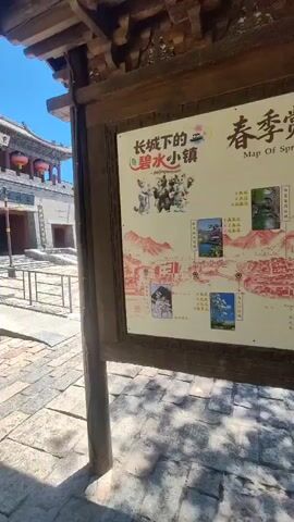 The video shows a sign with a map of spring flower viewing routes in a town called "Changshui Xiaozhen" (长水小镇). The sign is positioned outdoors, under a wooden awning, next to a stone pathway. The map itself is colorful, featuring illustrations of people and mountains, along with various photos of flowers at different locations marked on the map. The surrounding environment includes trees with dappled sunlight on the ground, and stone paving. In the background, a few people are visible, one person is sitting on a bench near the sign, and another is walking away further down the path. There is also a stone wall covered in ivy in the distance. The scene appears to be a tourist information point in a scenic town.
