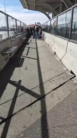 The video shows a walkway at an airport or similar transportation hub. The walkway has concrete barriers on either side, topped with glass panels and metal handrails. Benches are attached to the barriers. A group of people are walking along the walkway away from the camera, pulling luggage. The walkway surface is cracked and marked. Overhead is a metal awning. Shadows from the awning structure are visible on the walkway. In the background, aircraft and service vehicles can be seen through the glass panels. The sky is blue with some clouds.