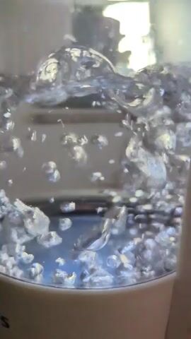 The video shows a close-up view of water boiling in a clear container, likely a kettle. Bubbles are rapidly forming and rising to the surface, creating turbulence and splashes. The water is bright and reflects light, giving it a shimmering appearance. The bottom of the container is visible, and it appears to be made of a white or cream-colored material. The water is clear and appears to be clean. The overall impression is one of vigorous heating and the energetic movement of water.