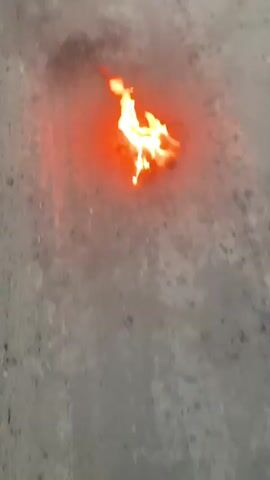 A close-up, top-down shot shows a small pile of something burning intensely on a grey, rough surface. The flames are bright yellow and orange, emitting a reddish glow that illuminates the immediate surroundings. The surface appears to be concrete or asphalt, with some scattered dark specks and a faint reddish hue spreading outwards from the fire. There are no people or other objects visible in the frame. The video is approximately 4 seconds long and shows no discernible change in the fire or its environment.
