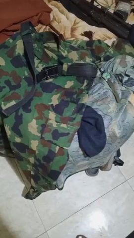 The video shows a close-up view of items laid out on a bed with a floral patterned bedspread. The primary focus is on a camouflage military uniform, complete with a dark belt and attached shoulder strap. Next to the uniform is a dark blue beanie and what appears to be a wristwatch. Lying across the top of the bed, extending into the upper right corner, is a black rifle with a strap. A brown garment is visible on the left side of the bed. Part of a person's foot is briefly visible at the bottom of the frame. The camera moves slightly during the video, providing different angles of the items on the bed. The environment appears to be indoors, likely a bedroom.