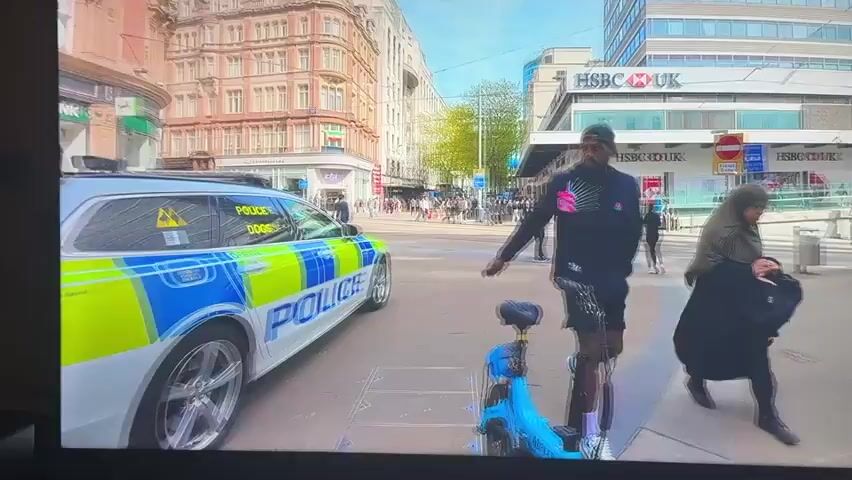 The video shows a street scene with a police car in the foreground. The police car has blue and yellow markings and the word "POLICE" written on it. A man in a police uniform is standing next to the car. There are other people walking in the background, as well as buildings and shops. The HSBC bank and Lloyds Bank are visible. There is also a bicycle parked near the police car. The scene appears to be a city street on a sunny day.