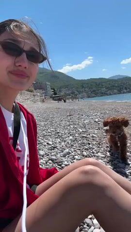 A bright and sunny daytime scene unfolds on a pebble beach in Zankovići, Montenegro. A young woman, wearing cat-eye sunglasses and a red zip-up hoodie over a white t-shirt with partial visible text "COLLE," is the main subject. Her dark hair is pulled back, and she smiles towards the camera initially. Beside her, a fluffy, brown dog, likely a poodle or similar breed, is visible.

The video captures an interaction between the woman and her dog. She is seen sitting on the pebbly shore, then turns and throws a small stick or twig into the distance, prompting the dog to enthusiastically run after it.

The background features a clear blue sky with the sun shining brightly, casting a warm glow. A vast expanse of calm, blue water stretches out, meeting the pebble beach. Along the coastline, various buildings are visible, backed by verdant, rolling mountains under the bright sky. The overall atmosphere is one of relaxation and enjoyment on a beautiful day.