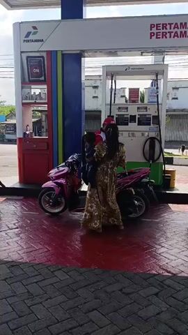 The video shows a woman holding a child while standing next to two pink motorbikes at a Pertamina gas station. The woman is wearing a long, patterned dress. A gas pump with a digital display and hoses is visible behind them. The ground is paved with bricks, and there is a red, wet-looking surface in front of the pump. Other vehicles and buildings are visible in the background. The scene appears to be during daylight.
