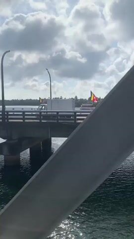 The video shows a bridge over a body of water on a cloudy day. There are several flags on the bridge, which appear to be religious or national flags. A white truck is driving across the bridge, and there are trees in the background. The water is dark and choppy, with sunlight reflecting on the surface. The bridge is made of concrete and has a railing. There are two lampposts on the bridge.