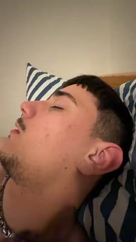 The video depicts a close-up of a young man sleeping on a bed. He is lying on his side with his head on a pillow that has blue and white stripes. He has short dark hair with a blunt fringe and a light beard. He is wearing a silver chain necklace. A hand comes into the frame and pokes his ear. The man wakes up slightly, yawns, and then closes his eyes again. The background is a plain white wall. The lighting is dim, suggesting it may be nighttime or a room with poor lighting.