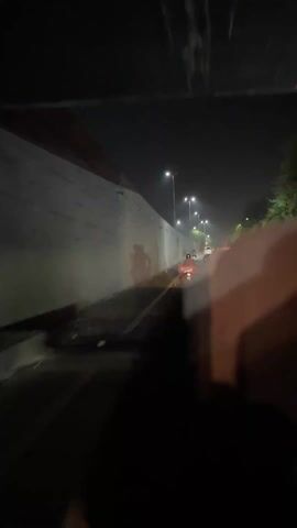 The video captures a night scene on a road, likely in an underpass or a section bordered by a high concrete wall on the left. Numerous vehicles, predominantly motorcycles, are seen traveling along the illuminated road. Several streetlights line the road, casting light and shadows on the wall and the moving traffic. A white line divides the road. The perspective is from a moving vehicle, suggesting the recording is taking place while driving. The setting is at night in Model Town, Pakistan. No discernible text is visible.