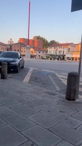The video shows a street scene in a town with buildings on either side. The street is paved with cobblestones in the foreground and asphalt in the background. There are several cars parked on the side of the street, including a black sedan. There is also a disabled parking sign visible. The sky is clear and blue, and the sun appears to be setting, casting a warm glow on the buildings. There are a few people walking in the background, but they are not clearly visible. The overall impression is one of a peaceful, quiet afternoon in a European town.