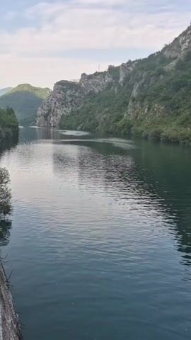 The video pans across a serene river with calm, turquoise water. The river is flanked by steep, rocky cliffs covered in lush green vegetation. The sky above is partly cloudy, with a soft, diffused light casting gentle reflections on the water's surface. The scene evokes a sense of natural beauty and tranquility.
