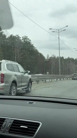 The video, filmed from inside a moving vehicle, shows the aftermath of a car accident on a highway. The sky is overcast, and water droplets are visible on the windshield, suggesting rain.

The primary subject is a dark gray Mitsubishi Lancer sedan, heavily damaged at its rear, parked on the left shoulder of the road. Its rear bumper is severely crunched and partially detached on the driver's side, where the taillight is also broken. The trunk lid is crumpled, and the rear windshield is shattered. The license plate "P 765 AM 96" is clearly visible on the back of the car. Road debris is scattered on the asphalt next to the damaged vehicle.

In the foreground, a blue SUV is visible on the right side of the frame. In the background, a dense forest lines the side of the road, separated by a metal guardrail. Several distinctive streetlights with multiple lamp heads are spaced along the road. A white semi-truck can be seen driving in the distance in the later part of the clip. The camera vehicle is driving past the scene.
