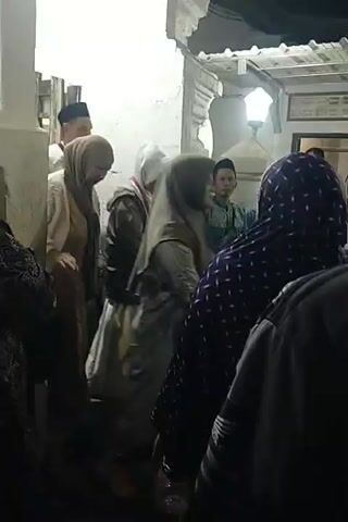 A group of people, some wearing Islamic attire like hijabs and skullcaps, are gathered in a dimly lit outdoor area that appears to be near a building with white walls and a traditional architectural style. The lighting suggests it is nighttime. In the foreground, several individuals are seen from behind, their clothing mostly dark. In the middle ground, a person in a light-colored jacket and hijab is walking, and another person in a light-colored shirt and skullcap is walking in the same direction. To the right, other individuals are visible, including a person in a dark purple headscarf with a patterned design. The overall atmosphere seems to be one of a gathering or an event.