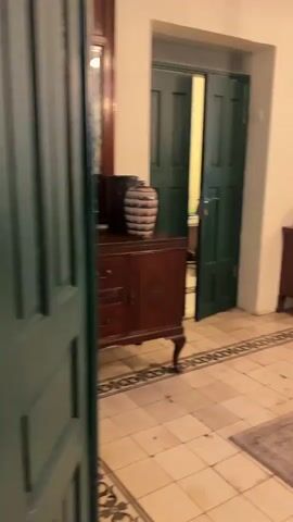 The video pans across a dimly lit, interior space. In the foreground, a large, rough-hewn wooden table with a glass top is visible. Beyond the table, a brown leather sofa, a grandfather clock, and a plant in a pot are situated against a wall. To the left, a closed dark green door is partially open, revealing a sliver of another room. Further left, a vintage piano with a stack of books and framed pictures on its surface is prominent. The floor is tiled with a decorative border. The walls are painted a pale color and adorned with framed pictures. The lighting appears to be a mix of ambient and artificial sources, creating a warm and somewhat vintage atmosphere. The video is a slow, panning motion, offering glimpses of various furniture and decorative items within the room.