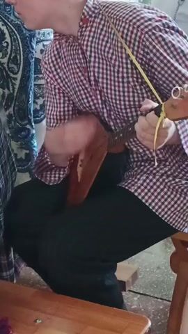 A young person is sitting on a wooden stool playing a balalaika. They are wearing a red and white checkered shirt, black pants, and have a yellow strap around their neck. The balalaika is brown, triangular, and has three strings. The person is strumming the instrument with their right hand and holding the neck with their left hand. There is a table to the left of the person, a curtain hanging behind them, and a wooden floor beneath them.