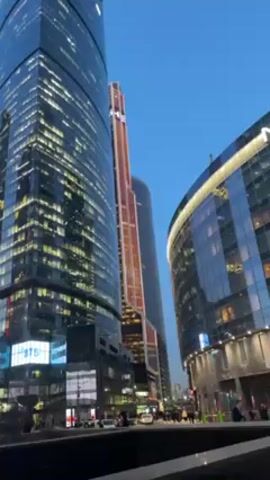 The video shows a low-angle view of several tall, modern buildings in an urban setting, likely a business district, against a clear blue sky at dusk or dawn. The buildings are primarily constructed of glass and steel, with many illuminated windows suggesting activity inside. One distinctive building is a red-toned skyscraper with a unique design element at its top.  The camera is stable, and the scene captures the scale and architectural characteristics of the area. A small group of people can be seen walking on the ground level between the buildings, and there are a few cars visible on the street. A sign for VTB Bank is visible on one of the lower buildings.  The general impression is of a contemporary and bustling urban environment.