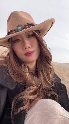 A woman with long brown hair and a cowboy hat is taking a selfie in a desert landscape. She's wearing a black coat and a light-colored dress or wrap. The background shows a sand dune with many people walking and sitting on it. A small lake can be seen in the distance. The woman is holding a small pink object in her hand. She makes different facial expressions for the selfie, including smiling and posing with her mouth slightly open. The scene appears to be taken during the day, under an overcast sky.
