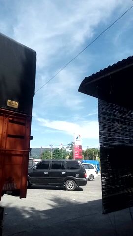 This media file captures a scene at a roadside area in Sukamulia, Indonesia. The perspective is low, looking upwards towards a bright blue sky with wispy clouds. In the foreground, the side of a large orange and black truck or cargo container is visible, its surface textured and worn.

The mid-ground of the scene features a paved area with several vehicles. A black SUV is parked, and a white car is also present. A prominent red and white sign for Pertamina, an Indonesian state-owned oil and natural gas corporation, stands tall, indicating a gas station is nearby. Green trees and vegetation are visible behind the vehicles, suggesting a somewhat rural or suburban setting.

In the foreground, a dark structure, possibly an awning or roof with vertical slats, creates a framed view of the scene beyond. The light suggests it is daytime, likely late morning or early afternoon, given the bright sunlight and clear sky. The overall atmosphere appears casual and ordinary, depicting a typical day in the described location.

Visible text includes "PERTAMINA" on the gas station sign. There is also some text on the side of the orange container in the foreground, but it is largely obscured and difficult to read. The image conveys a sense of everyday life, with vehicles and infrastructure common to the region.