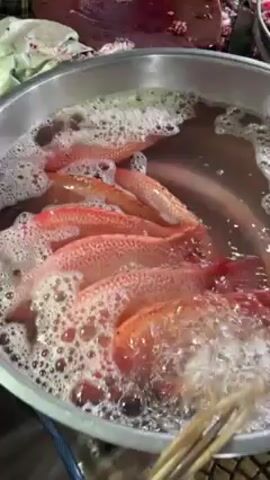 The video shows a metal bowl filled with several small, red fish submerged in bubbly water. A wooden skewer rests in the bowl alongside the fish, which appear to be undergoing some sort of cleaning or preparation process. To the left of the bowl, there is a wooden cutting board with a large piece of meat on it, and other unidentified items in green plastic wrapping. To the right of the bowl, another metal bowl contains what appears to be raw meat. The overall scene suggests a food preparation area, possibly a market or kitchen.