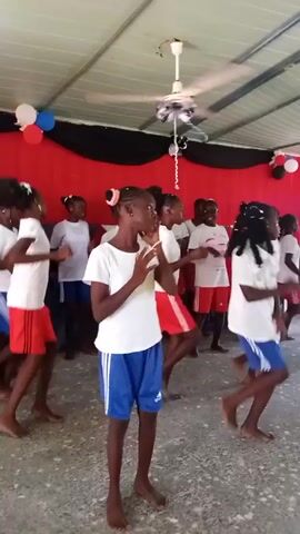 A group of young African girls are performing a choreographed dance routine in what appears to be an indoor or covered outdoor space. The girls are barefoot and wearing matching white t-shirts. Some have red shorts, while others have blue shorts with white stripes down the side. There are about 15 girls, and they are spread out in a loose formation.  Behind them is a red and black backdrop, decorated with balloons in red, white, and blue. A ceiling fan is visible above them. The room is well lit and has a concrete floor.