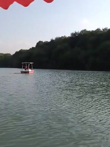 The video shows a view of a lake with a small white and red boat in the middle of it. The boat has a canopy and appears to have people inside. The lake is surrounded by a dense forest with trees. The sky is visible and appears to be clear. There is a red canopy in the foreground of the video, suggesting the footage is being filmed from another boat. Another boat is visible in the distance on the left side of the screen. The water is calm and rippling.