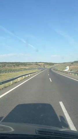 The video shows a car's perspective while driving on a highway under a clear blue sky. The road stretches into the distance with sparse vegetation on either side. Guardrails line the highway.
