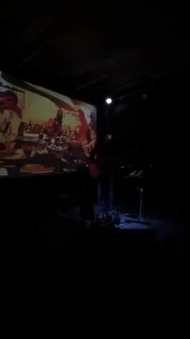 A musician plays guitar on a dark stage in front of a projected screen. The screen displays “PABLO MILLER” in white text, overlaid on a collage of colors and images, including what appears to be a hand holding a musical instrument. A microphone stand and keyboard stand are visible near the musician, who is dressed in dark clothing. The stage is dimly lit by a single light above and some smaller stage lights. The atmosphere suggests a live music performance.
