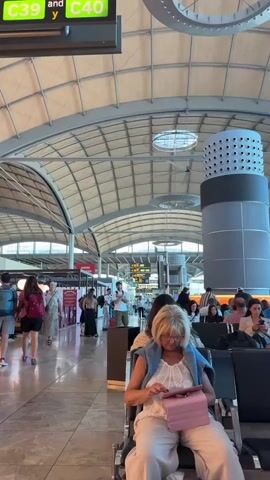 The video shows the interior of an airport terminal. People are seen walking through the terminal in the background, some with luggage. The space has a high, arched ceiling with a metallic frame and a series of round, decorative light fixtures hanging from it. There's a large, cylindrical structure with a perforated pattern. 
In the foreground, an older woman is sitting on a black chair, using a pink tablet. She is wearing a white top, light-colored pants, and a blue sweater draped over her shoulders. A sign overhead indicates gates C39 and C40.