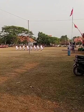 A group of young people in white uniforms are marching in formation across a grassy field. They are accompanied by two adults, one of whom is holding a flag. There are buildings and trees in the background, and a motorcycle is parked in the foreground. The sky is clear and the lighting suggests it is daytime.