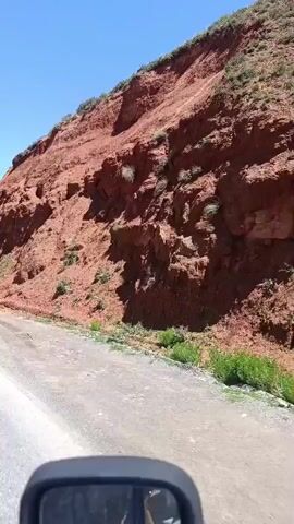 The video shows a red rock cliff face next to a dirt road. The cliff is steep and appears eroded, with some sparse vegetation growing on it. The road is in the foreground, and a car's side mirror is visible at the bottom of the frame. The sky is clear and blue, indicating a sunny day. The camera is mounted on a moving vehicle, suggesting the video was taken from a car driving along the road.