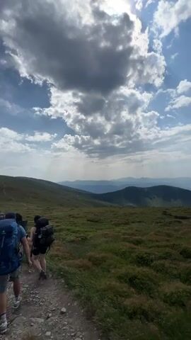 The video begins with a wide shot of a grassy mountainous landscape under a cloudy sky. The camera then pans to show more of the rolling hills and a distant mountain peak. The final clips focus on a young woman with brown hair, wearing a plaid shirt and a large backpack, standing on a rocky trail in the mountains. She is holding a hiking pole and looking out at the scenic view. The environment is characterized by steep, green mountains covered in grass and shrubbery, with a clear blue sky in the background.
