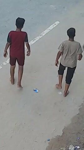This is an aerial video of a group of young boys playing in an outdoor area. The ground appears to be asphalt or concrete, with a white line painted across it. In the foreground, two boys are walking away from the camera. The boy on the left is wearing a red t-shirt and shorts. The boy on the right is wearing a dark t-shirt and dark pants. Further back, other boys can be seen walking and interacting. One boy in a green t-shirt is walking away, and another boy in a black t-shirt is standing. There is a large object that appears to be a rope or cable hanging from the top right of the frame, partially obscuring the view. A football is visible in the background. The overall impression is of a casual outdoor game among children.