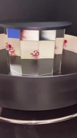 A video displays a rotating cylindrical object with multiple mirrored surfaces. Each surface features a red stick figure in a running pose. The figures are positioned facing outwards and appear to be in mid-stride. The cylinder is rotating on a black base, and reflections of the figures are visible on the mirrored surfaces. Above the cylinder is a black circular top. The background is dark.