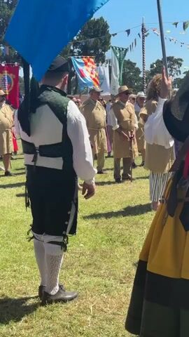 A group of people are standing in a field. The person in the foreground on the left is wearing a dark green vest with white trim over a white shirt. They also have on white socks with a patterned design and dark shoes. They are holding bagpipes. In the foreground on the right, a person is wearing a yellow skirt with a dark hem and a brown top with red straps. Behind them, a line of people are dressed in tan tunics. Some are wearing hats. There are flags and banners visible in the background, and trees and a blue sky are overhead. The ground is covered in green grass.