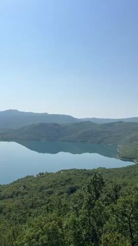 The video shows a wide, panoramic view of a large lake surrounded by lush green hills and mountains. The sky is clear and blue. In the foreground, there is a dense forest. As the video progresses, the camera pans slightly, revealing more of the lake and the landscape. Several small islands covered in vegetation can be seen dotting the water. The overall impression is one of a serene and natural landscape. There are no people or man-made structures visible in the video.