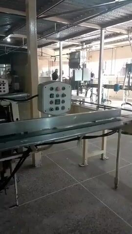 The video displays an industrial setting, likely a factory floor, with a conveyor belt system prominently featured. The environment appears to be a large, open space with a metal roof supported by industrial beams and pillars. Natural light filters in from windows in the background, illuminating the scene. The foreground shows a portion of a conveyor belt made of metal, with a control panel nearby. This panel has several buttons and indicators. Further into the scene, other parts of the conveyor system are visible, along with what appear to be processing machines and some equipment suspended from the ceiling. In the background, two individuals can be seen in silhouette, seemingly engaged in work or moving through the facility. One person is standing near some machinery, while the other is in the process of walking. The overall impression is one of an active, if somewhat dated, manufacturing environment.