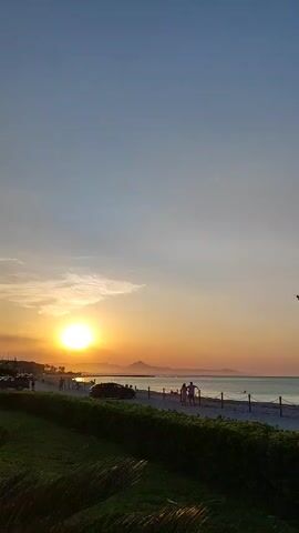 This is a landscape video showing a beach scene during sunset. The sun is visible on the left side of the frame, casting a warm glow over the water and sky. The ocean stretches out to the horizon, with a few boats visible in the distance. A sandy beach runs along the coastline, bordered by a low green hedge in the foreground. There are a few people walking along the beach and a car parked nearby. The sky is a gradient of colors, from blue at the top to orange and yellow near the horizon, indicating the sun is setting.