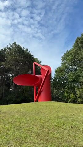 A large, abstract red sculpture stands on a grassy hill. The sculpture is composed of several geometric shapes, including a large funnel-like form extending to the left, a cylindrical base, and a triangular element connecting the two. The background consists of green trees and a blue sky with scattered clouds. The camera slowly pans to the right, revealing more of the sculpture and the surrounding landscape. The lighting suggests it is daytime, possibly late morning or early afternoon. No people are visible in the video.