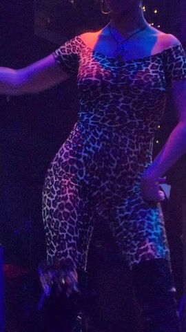 The video shows a woman dancing on a stage, wearing a tight-fitting leopard print jumpsuit and black knee-high boots. The stage has multicolored lights shining on her, alternating between blues, greens, yellows, and oranges. The camera angle is low, looking up at the dancer. In the foreground, the top of someone's head is visible, wearing a dark hat or bandana with a paisley-like pattern.
