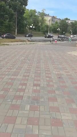 The video shows a paved walkway with red and gray bricks. In the background, there is a road with several cars driving by. Trees line the road, and in the distance, there are buildings. Several people are visible walking on the sidewalk or near the road. The sky is blue with some clouds. The camera is panning slowly to the right.
