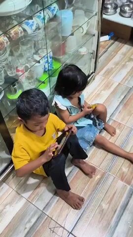 Two young children sit cross-legged on a tiled floor in front of a glass-fronted cabinet. The boy, wearing a yellow shirt and black pants, is engrossed in a tablet. The girl, in a light blue top and denim shorts, is focused on what appears to be a small object in her hands, possibly drawing with a pencil. The cabinet behind them is filled with various glass items, including plates, cups, and bowls. The floor tiles are a mix of beige and brown, resembling wood planks.