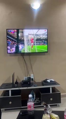 The video shows a room with a television mounted on a beige wall, displaying a Premier League soccer match between Chelsea (CHE) and Nottingham Forest (NFO). The score is 1-1. A black TV stand is beneath the television, holding a laptop, a PlayStation 4, a controller, a small fan, and an iron, along with various cables. On the coffee table, in the foreground, there is a bottle of water, a cell phone, and a plastic bag filled with groceries. Above the TV, there is a single light fixture. The camera remains stationary.