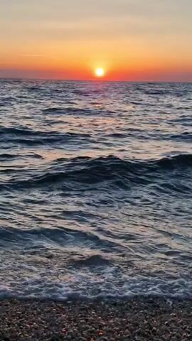 The video captures a serene sunset over the Adriatic Sea in Zadar, Croatia. The sun, a radiant orange orb, is low on the horizon, casting a warm glow across the sky and reflecting shimmering paths on the water's surface. Gentle waves lap at a pebbled shore in the foreground. The water itself is a deep blue, with lighter, foamy crests as the waves break. The sky transitions from a soft orange near the sun to a pale yellow and then a muted pinkish hue higher up. There are no people or objects visible in the shot, emphasizing the natural beauty of the scene. The overall mood is peaceful and tranquil, inviting contemplation of the day's end. The time of day is clearly sunset, and the weather appears to be calm with clear skies. The presence of the sea and pebbles suggest a coastal location. No text is visible in the media file.